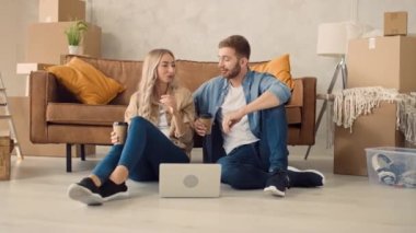 Couple in a New Home Using Laptop Drinking Coffee. Couple Using Laptop for Shopping, Booking Tickets, Discussing Interior. Girl Pointing in Screen.