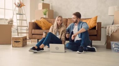 Couple in a New Home Using Laptop Drinking Coffee. Couple Using Laptop for Shopping, Booking Tickets, Discussing Interior. Girl Pointing in Screen.