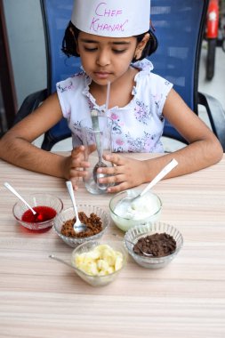 Tatlı Hintli aşçı kız vanilyalı dondurma, browni, kakao tozu, taze doğranmış meyve ve çilek şurubu içeren ateşte pişirilmemiş dondurma yemeğinin bir parçası olarak sundae hazırlıyor. Küçük çocuk yemek hazırlıyor.