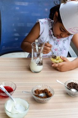 Tatlı Hintli aşçı kız vanilyalı dondurma, browni, kakao tozu, taze doğranmış meyve ve çilek şurubu içeren ateşte pişirilmemiş dondurma yemeğinin bir parçası olarak sundae hazırlıyor. Küçük çocuk yemek hazırlıyor.