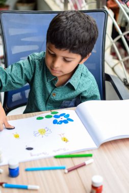 Zeki Hintli küçük çocuk yaz tatillerinde farklı renklerde su rengi setiyle baş parmak boyası yapıyor, şirin Hintli çocuk ahşap masaya renkli başparmak resimleri çiziyor.