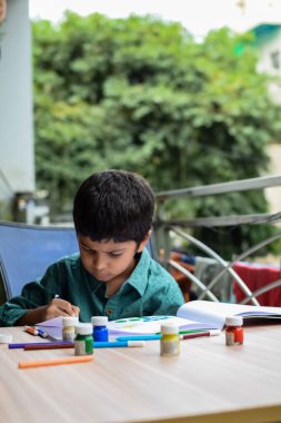 Zeki Hintli küçük çocuk yaz tatillerinde farklı renklerde su rengi setiyle baş parmak boyası yapıyor, şirin Hintli çocuk ahşap masaya renkli başparmak resimleri çiziyor.