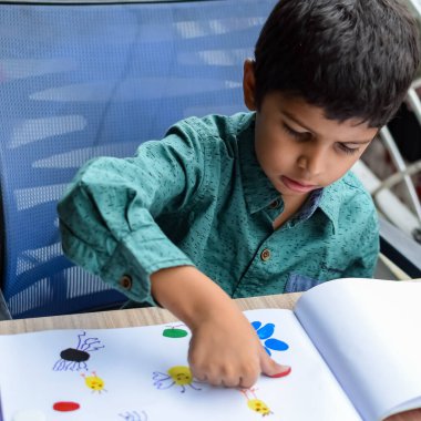 Zeki Hintli küçük çocuk yaz tatillerinde farklı renklerde su rengi setiyle baş parmak boyası yapıyor, şirin Hintli çocuk ahşap masaya renkli başparmak resimleri çiziyor.