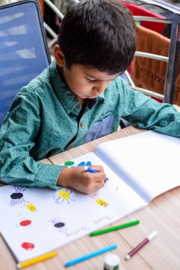 Zeki Hintli küçük çocuk yaz tatillerinde farklı renklerde su rengi setiyle baş parmak boyası yapıyor, şirin Hintli çocuk ahşap masaya renkli başparmak resimleri çiziyor.