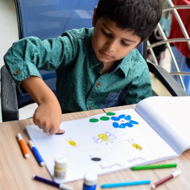 Zeki Hintli küçük çocuk yaz tatillerinde farklı renklerde su rengi setiyle baş parmak boyası yapıyor, şirin Hintli çocuk ahşap masaya renkli başparmak resimleri çiziyor.