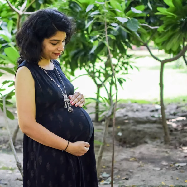 A pregnant Indian lady poses for outdoor pregnancy shoot and hands on belly, Indian pregnant woman puts her hand on her stomach with a maternity dress at society park, Pregnant outside maternity shoot