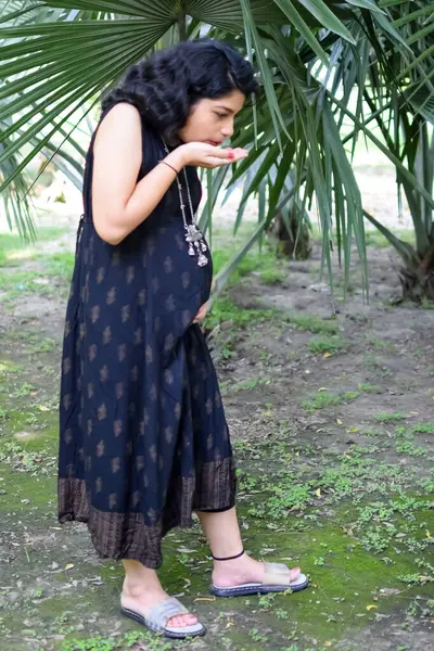 A pregnant Indian lady poses for outdoor pregnancy shoot and hands on belly, Indian pregnant woman puts her hand on her stomach with a maternity dress at society park, Pregnant outside maternity shoot