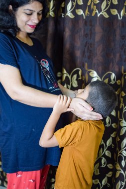 Hamile bir Hintli kadın çocuğunu evde doğurmak için poz veriyor ve ellerini karnının üstüne koyuyor. Hintli hamile kadın elini karnına koyuyor ve evinde hamile elbisesi var.