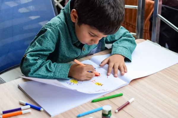 Zeki Hintli küçük çocuk yaz tatillerinde farklı renklerde su rengi setiyle baş parmak boyası yapıyor, şirin Hintli çocuk ahşap masaya renkli başparmak resimleri çiziyor.