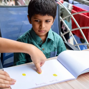 Zeki Hintli küçük çocuk yaz tatillerinde farklı renklerde su rengi setiyle baş parmak boyası yapıyor, şirin Hintli çocuk ahşap masaya renkli başparmak resimleri çiziyor.