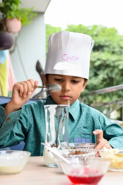 Cute Indian chef boy preparing sundae dish as a part of non fire cooking which includes vanilla ice cream, brownie, coco powder, freshly chopped fruits and strawberry syrup. Little kid preparing food
