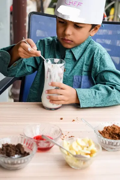 Cute Indian chef boy preparing sundae dish as a part of non fire cooking which includes vanilla ice cream, brownie, coco powder, freshly chopped fruits and strawberry syrup. Little kid preparing food