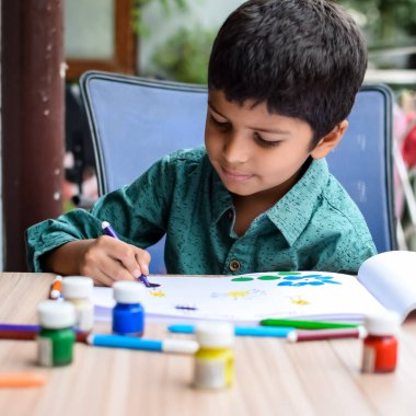 Zeki Hintli küçük çocuk yaz tatillerinde farklı renklerde su rengi setiyle baş parmak boyası yapıyor, şirin Hintli çocuk ahşap masaya renkli başparmak resimleri çiziyor.