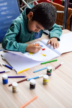 Zeki Hintli küçük çocuk yaz tatillerinde farklı renklerde su rengi setiyle baş parmak boyası yapıyor, şirin Hintli çocuk ahşap masaya renkli başparmak resimleri çiziyor.