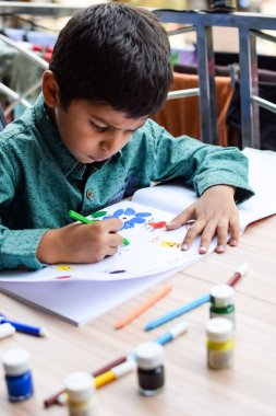 Zeki Hintli küçük çocuk yaz tatillerinde farklı renklerde su rengi setiyle baş parmak boyası yapıyor, şirin Hintli çocuk ahşap masaya renkli başparmak resimleri çiziyor.