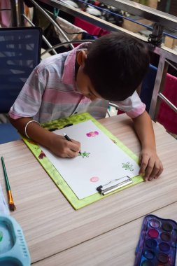 Zeki Hintli küçük çocuk yaz tatillerinde farklı renklerde su rengi setiyle baş parmak boyası yapıyor, şirin Hintli çocuk ahşap masaya renkli başparmak resimleri çiziyor.