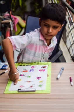 Zeki Hintli küçük çocuk yaz tatillerinde farklı renklerde su rengi setiyle baş parmak boyası yapıyor, şirin Hintli çocuk ahşap masaya renkli başparmak resimleri çiziyor.