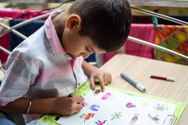 Zeki Hintli küçük çocuk yaz tatillerinde farklı renklerde su rengi setiyle baş parmak boyası yapıyor, şirin Hintli çocuk ahşap masaya renkli başparmak resimleri çiziyor.