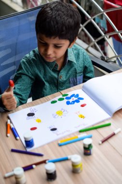 Zeki Hintli küçük çocuk yaz tatillerinde farklı renklerde su rengi setiyle baş parmak boyası yapıyor, şirin Hintli çocuk ahşap masaya renkli başparmak resimleri çiziyor.