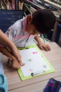 Zeki Hintli küçük çocuk yaz tatillerinde farklı renklerde su rengi setiyle baş parmak boyası yapıyor, şirin Hintli çocuk ahşap masaya renkli başparmak resimleri çiziyor.