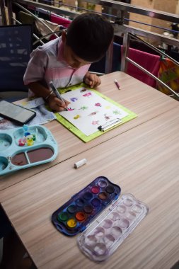 Zeki Hintli küçük çocuk yaz tatillerinde farklı renklerde su rengi setiyle baş parmak boyası yapıyor, şirin Hintli çocuk ahşap masaya renkli başparmak resimleri çiziyor.