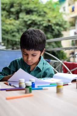 Zeki Hintli küçük çocuk yaz tatillerinde farklı renklerde su rengi setiyle baş parmak boyası yapıyor, şirin Hintli çocuk ahşap masaya renkli başparmak resimleri çiziyor.