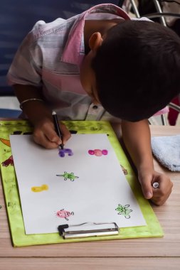 Zeki Hintli küçük çocuk yaz tatillerinde farklı renklerde su rengi setiyle baş parmak boyası yapıyor, şirin Hintli çocuk ahşap masaya renkli başparmak resimleri çiziyor.