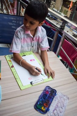 Zeki Hintli küçük çocuk yaz tatillerinde farklı renklerde su rengi setiyle baş parmak boyası yapıyor, şirin Hintli çocuk ahşap masaya renkli başparmak resimleri çiziyor.