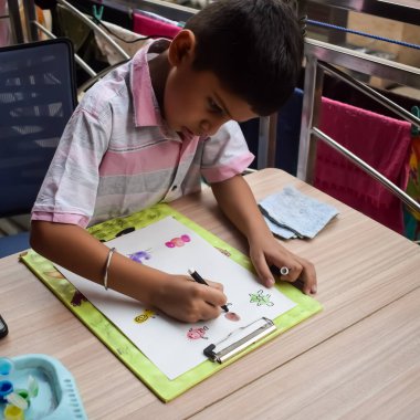 Zeki Hintli küçük çocuk yaz tatillerinde farklı renklerde su rengi setiyle baş parmak boyası yapıyor, şirin Hintli çocuk ahşap masaya renkli başparmak resimleri çiziyor.