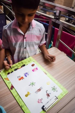 Zeki Hintli küçük çocuk yaz tatillerinde farklı renklerde su rengi setiyle baş parmak boyası yapıyor, şirin Hintli çocuk ahşap masaya renkli başparmak resimleri çiziyor.