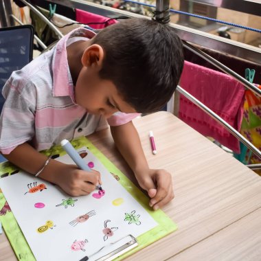 Zeki Hintli küçük çocuk yaz tatillerinde farklı renklerde su rengi setiyle baş parmak boyası yapıyor, şirin Hintli çocuk ahşap masaya renkli başparmak resimleri çiziyor.