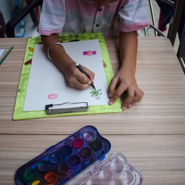 Zeki Hintli küçük çocuk yaz tatillerinde farklı renklerde su rengi setiyle baş parmak boyası yapıyor, şirin Hintli çocuk ahşap masaya renkli başparmak resimleri çiziyor.