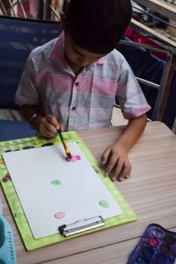 Zeki Hintli küçük çocuk yaz tatillerinde farklı renklerde su rengi setiyle baş parmak boyası yapıyor, şirin Hintli çocuk ahşap masaya renkli başparmak resimleri çiziyor.