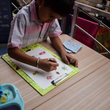 Zeki Hintli küçük çocuk yaz tatillerinde farklı renklerde su rengi setiyle baş parmak boyası yapıyor, şirin Hintli çocuk ahşap masaya renkli başparmak resimleri çiziyor.