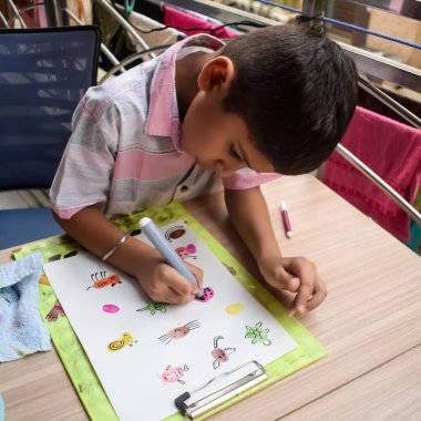 Zeki Hintli küçük çocuk yaz tatillerinde farklı renklerde su rengi setiyle baş parmak boyası yapıyor, şirin Hintli çocuk ahşap masaya renkli başparmak resimleri çiziyor.