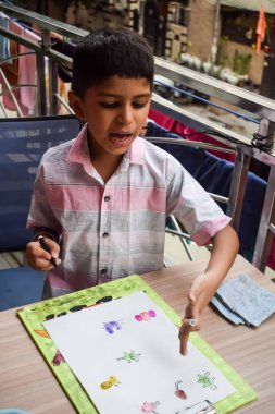 Zeki Hintli küçük çocuk yaz tatillerinde farklı renklerde su rengi setiyle baş parmak boyası yapıyor, şirin Hintli çocuk ahşap masaya renkli başparmak resimleri çiziyor.