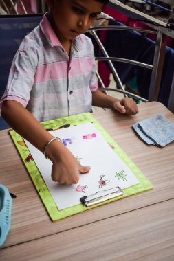 Zeki Hintli küçük çocuk yaz tatillerinde farklı renklerde su rengi setiyle baş parmak boyası yapıyor, şirin Hintli çocuk ahşap masaya renkli başparmak resimleri çiziyor.