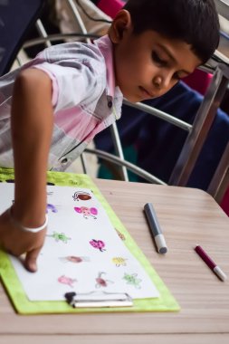 Zeki Hintli küçük çocuk yaz tatillerinde farklı renklerde su rengi setiyle baş parmak boyası yapıyor, şirin Hintli çocuk ahşap masaya renkli başparmak resimleri çiziyor.