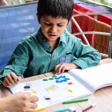 Zeki Hintli küçük çocuk yaz tatillerinde farklı renklerde su rengi setiyle baş parmak boyası yapıyor, şirin Hintli çocuk ahşap masaya renkli başparmak resimleri çiziyor.