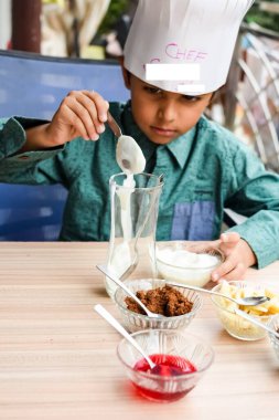 Tatlı Hintli aşçı çocuk vanilyalı dondurma, browni, kakao tozu, taze doğranmış meyve ve çilek şurubu içeren ateşte pişirilmemiş dondurma yemeğinin bir parçası olarak sundae hazırlıyor. Küçük çocuk yemek hazırlıyor.