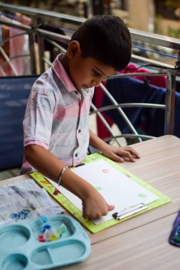 Zeki Hintli küçük çocuk yaz tatillerinde farklı renklerde su rengi setiyle baş parmak boyası yapıyor, şirin Hintli çocuk ahşap masaya renkli başparmak resimleri çiziyor.