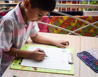 Zeki Hintli küçük çocuk yaz tatillerinde farklı renklerde su rengi setiyle baş parmak boyası yapıyor, şirin Hintli çocuk ahşap masaya renkli başparmak resimleri çiziyor.