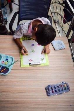 Zeki Hintli küçük çocuk yaz tatillerinde farklı renklerde su rengi setiyle baş parmak boyası yapıyor, şirin Hintli çocuk ahşap masaya renkli başparmak resimleri çiziyor.