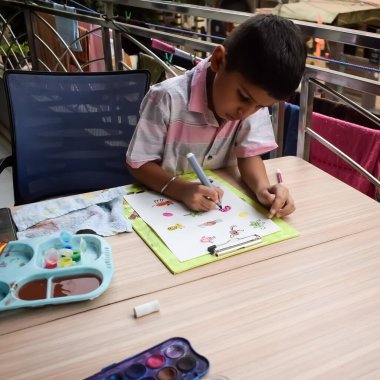 Zeki Hintli küçük çocuk yaz tatillerinde farklı renklerde su rengi setiyle baş parmak boyası yapıyor, şirin Hintli çocuk ahşap masaya renkli başparmak resimleri çiziyor.