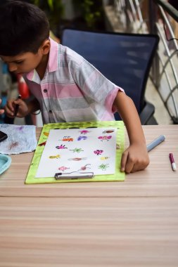 Zeki Hintli küçük çocuk yaz tatillerinde farklı renklerde su rengi setiyle baş parmak boyası yapıyor, şirin Hintli çocuk ahşap masaya renkli başparmak resimleri çiziyor.