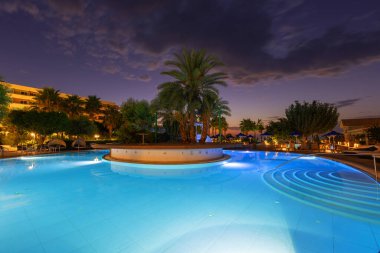 Şafak vakti, berrak bir su havuzu ve arka planda büyüyen palmiye ağaçları bulutlu ve yükselen güneşli güzel bir gökyüzü.