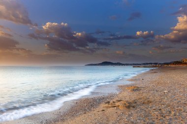 Güzel kumlu - gün doğumunda Rhodes adasında çakıl taşlı uzun plaj. Beyaz dalgalar kıyıya vuruyor. Arka planda mavi bir gökyüzü var..