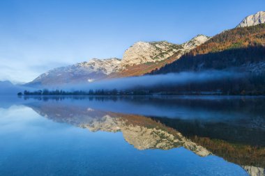 Avusturya Gölü Grundlsee, dağ zirvelerini yansıtan güzel, sakin bir göl yüzeyi. Sabah sisi yüzeyde yuvarlanıyor..