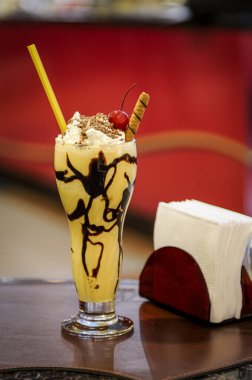 Milkshake with chocolate syrup, whipped cream and cherry, on a coffee shop table with blurred red background. clipart