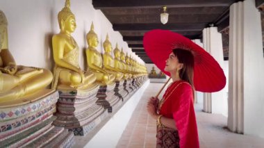 Tayland 'ın Wat Phutthaisawan, Ayutthaya' da dizilmiş birçok Buda heykeline hürmetlerini sunan, altınla süslenmiş kırmızı Tayland elbisesi giymiş genç bir kadın.